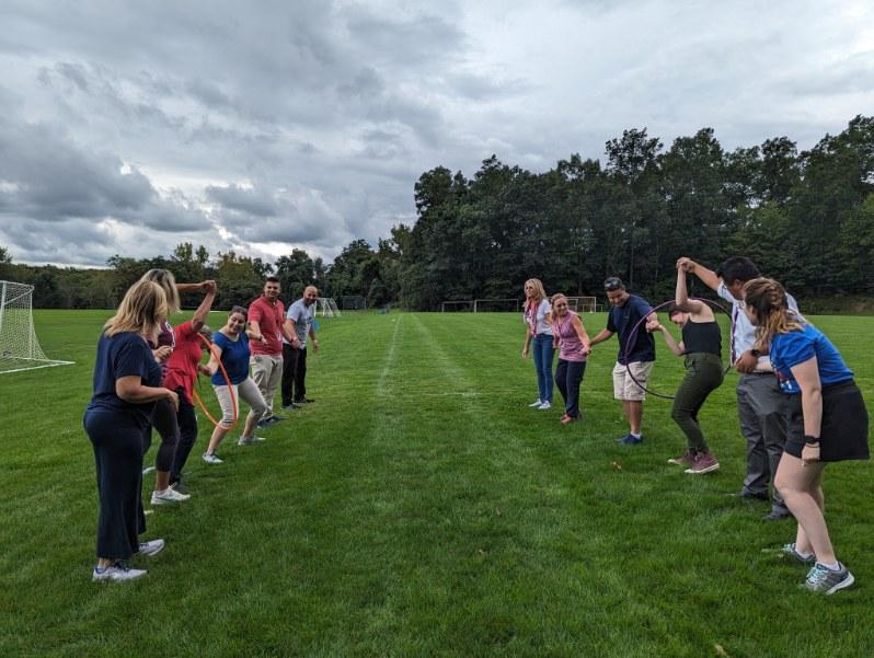 Two rows of adults stand together holding hands as they pass a hula hoop down the row.