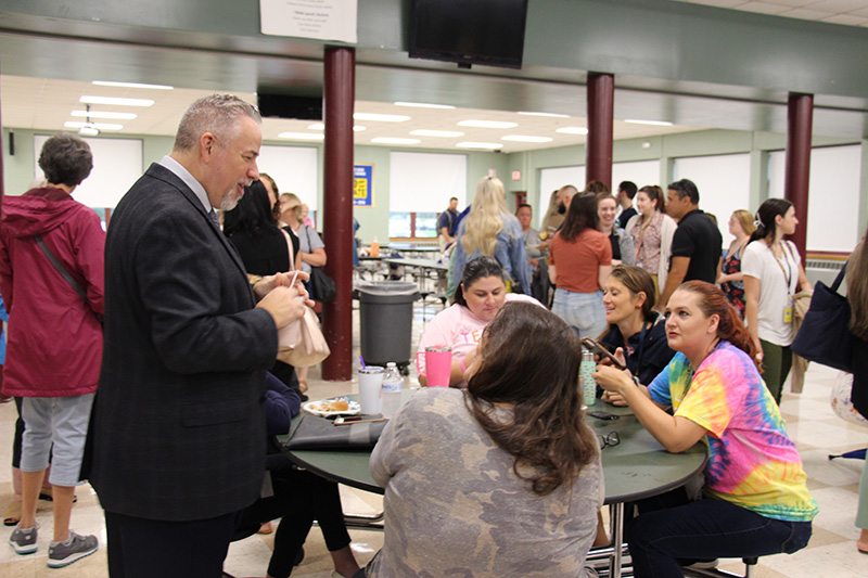 A man in a jacket and tie stands talking to a group of four women who are sitting at a table.