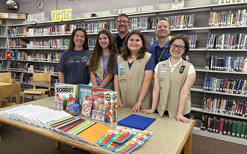 Three young women wearing beige girl scout uniforms smile and stand behind a table that has games and school supplies and playing cards. Next to them is a woman and behind them are two men. All are smiling.