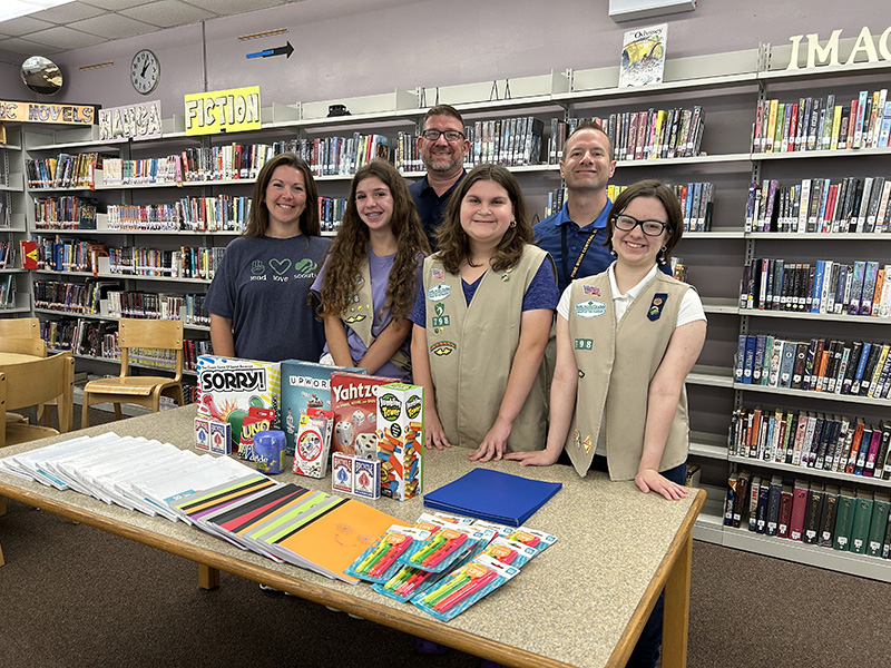 Three young women wearing beige girl scout uniforms smile and stand behind a table that has games and school supplies and playing cards. Next to them is a woman and behind them are two men. All are smiling.