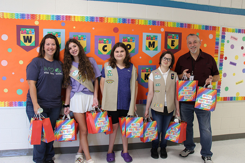 A woman on the left, three middle school girls in the center wearing beige girl scout vests, and a man on the right all hold birthday gift bags. They are all smiling.