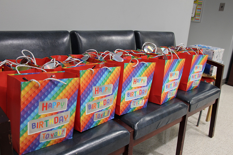 Twenty rainbow colored bags that say Happy Birthday on them sit on a bank of chairs.