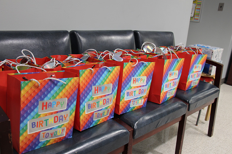 Twenty rainbow colored bags that say Happy Birthday on them sit on a bank of chairs.