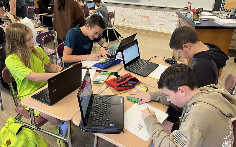 Four middle school boys work together at a group of four desks pushed together, writing on paper, all with their chromebooks open.