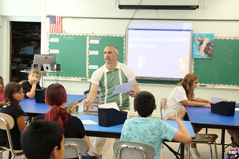 A man dressed in a green and cream shirt hands out papers to students sitting at tables.