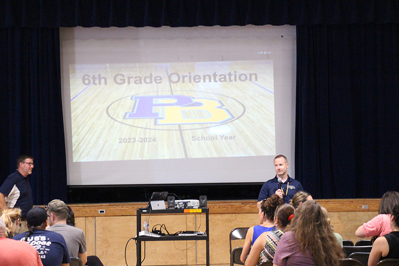 Two men at the front of a large gym in front of a group of adults. Behind them is a large screen with a slide on it that says 6th Grade Orientation with a large blue and gold PB on it.