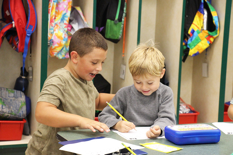 A third-grade boy sits next to a kindergarten boy who is holding a pencil. They are both smiling.