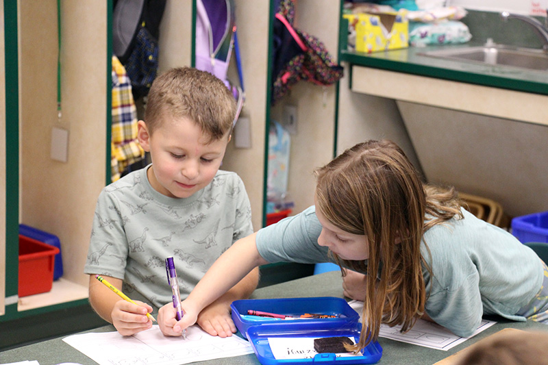 A third-grade girl reaches across a table to write on a piece of paper. A kindergarten boy holds a pencil and watches what she writes.