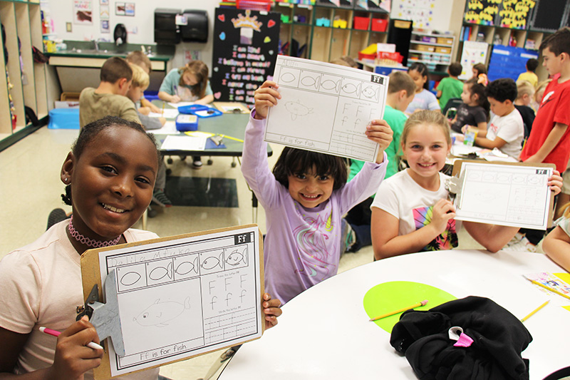 Three elementary age girls - two third-graders and a kindergartener in the center, hold up their worksheets and smile.