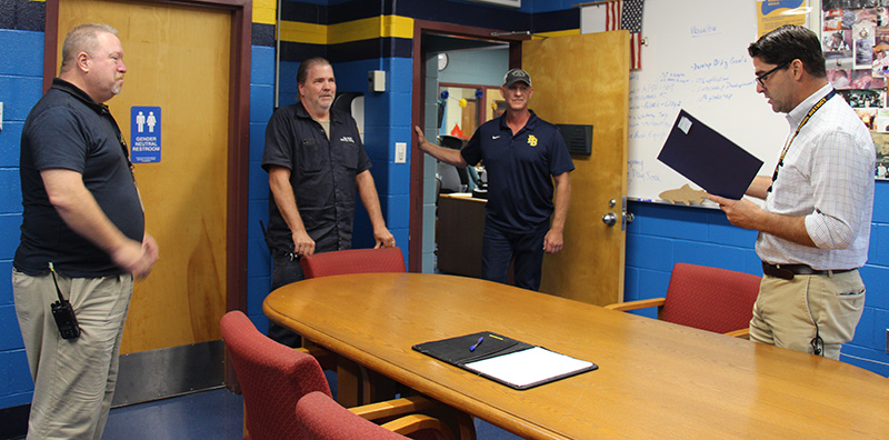 Four men stand around a table. the man on the right is reading from a folder as the other three listen.