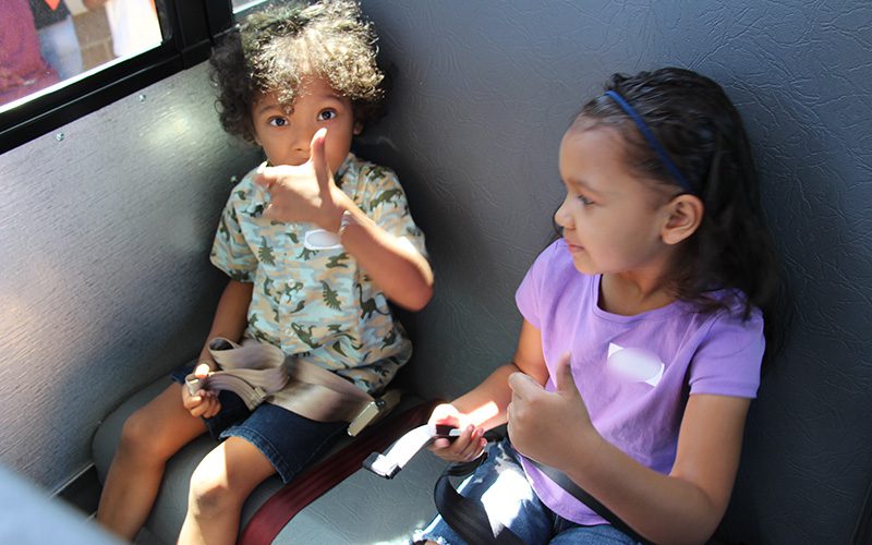 Two kindergarten kids sit in a big bus seat. They are both giving thumbs up.
