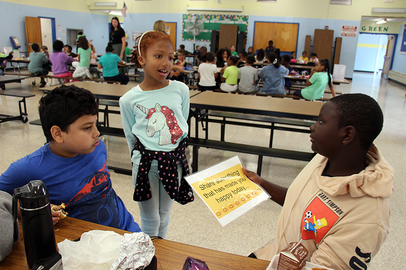 Two fourth-grade boys sit at a table. The boy on the right holds a piece of paper that says "Share something that has made you happy today" A girl stands there answering him.