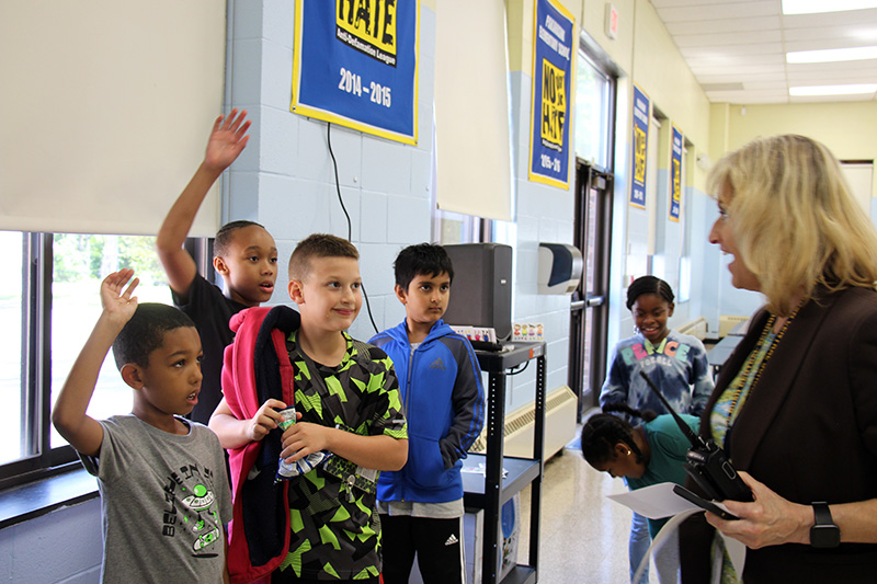 A group of four elementary age students stand talking to a woman who is smiling. Two of the boys have their hands up.