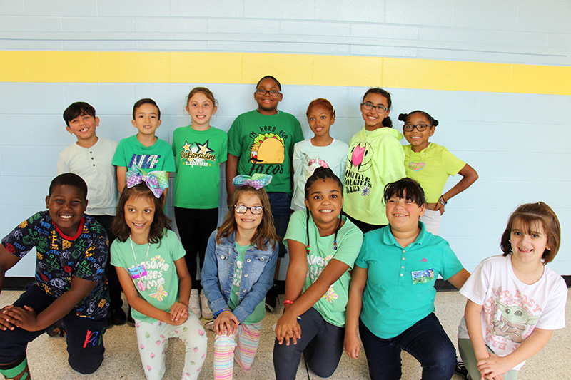 A group of 13 fourth-grade students, all wearing green, pose for a picture together. They are all smiling.