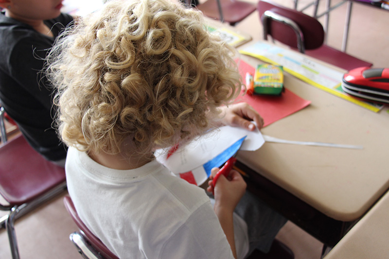 A third grade student with long curly blonde hair, cuts paper that is red white and blue.