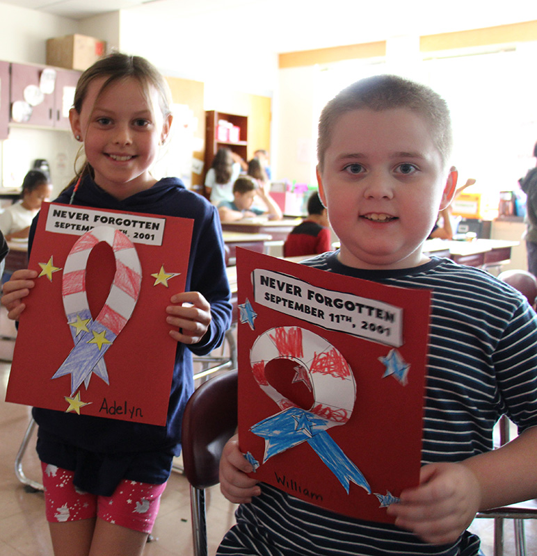 Two third-grade students - a girl on the left and a boy on the right - each hold a red piece of construction paper with a red, white and blue cut out ribbon they made.