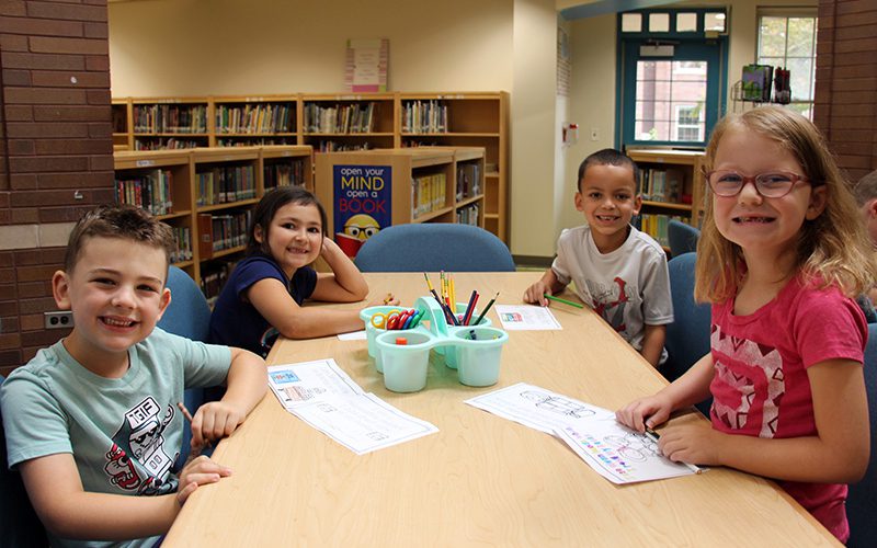 a group of four first-grade students color their books at a table. They are all looking up and smiling.