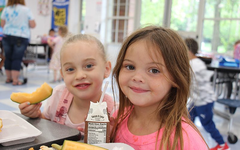 Two kindergarten girls eat lunch together and smile. One has chocolate milk in her hand and the other is holding a piece of cantaloupe.