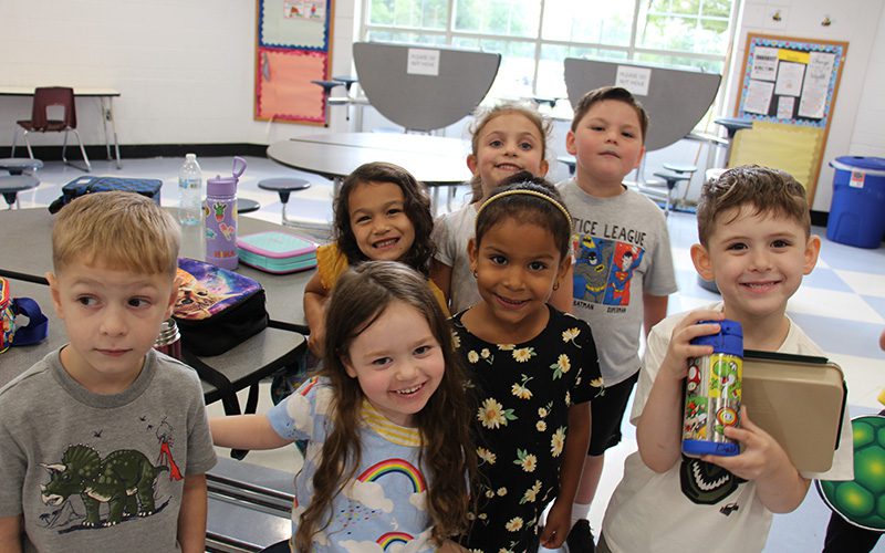 A group of seven kindergarten students in a cafeteria, all standing together smiing.