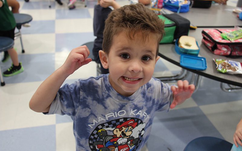 A young boy with a blue tie-dye t shirt on that says Mari Kart with a picture, He has short brow hair and is smiling