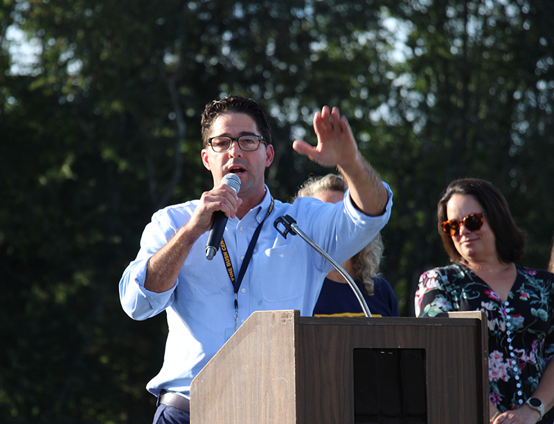 A man with dark hair and glasses speaks at a podium.