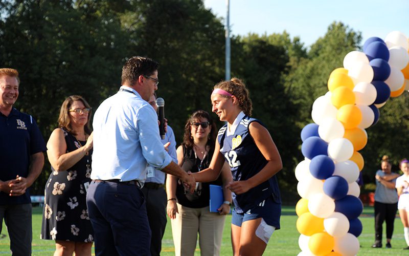 A man in a blue shirt and pants shakes the hand of a high school girl dressed in a soccer uniform.