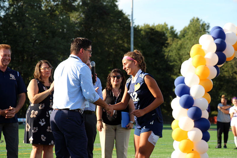 A man in a blue shirt and pants shakes the hand of a high school girl dressed in a soccer uniform.