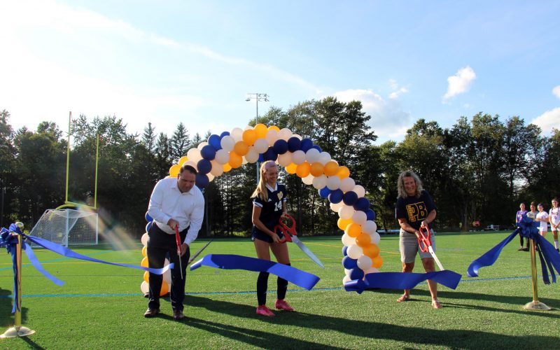 Three people hold oversized scissors and cut a blue ribbon on a field.