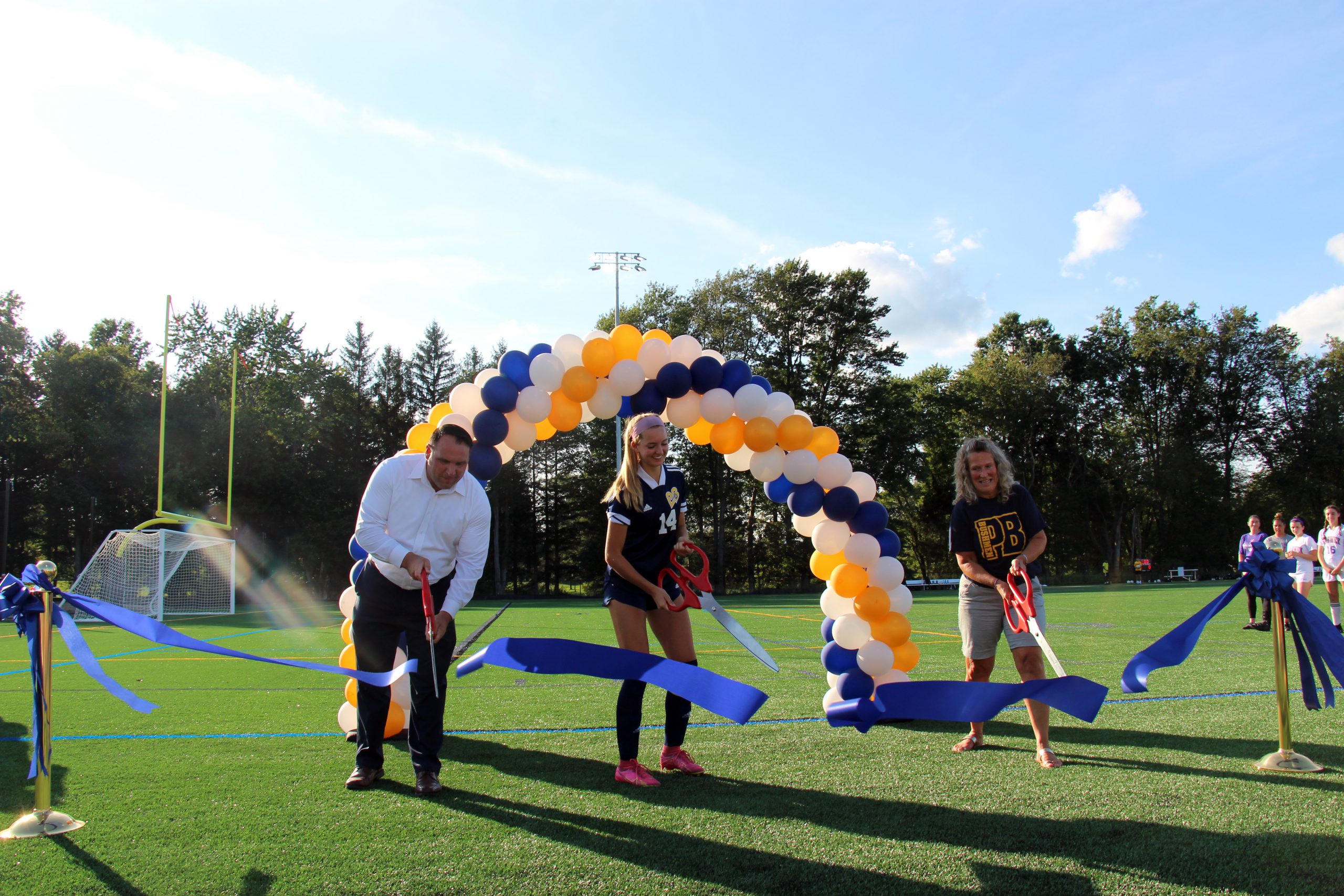 Three people hold oversized scissors and cut a blue ribbon on a field.