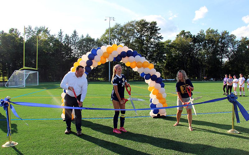 A beautiful sunny day with blue skies. Three people - a man on the left, a high school student in a blue and white soccer uniform in the center, and a woman on the right, stand by a large blue ribbon. Each has oversized scissors to cut it.