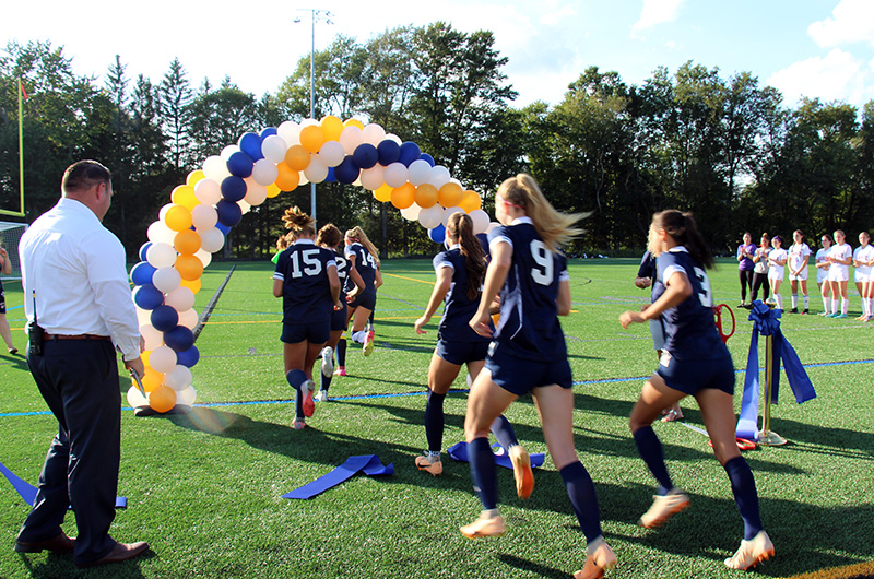A high school soccer team runs through a blue gold and white arch of balloons.