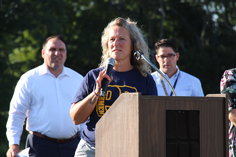 A woman with blonde hair, wearing a blue tshirt with a gold PB on it, speaks at a podium. There are two men behind her.