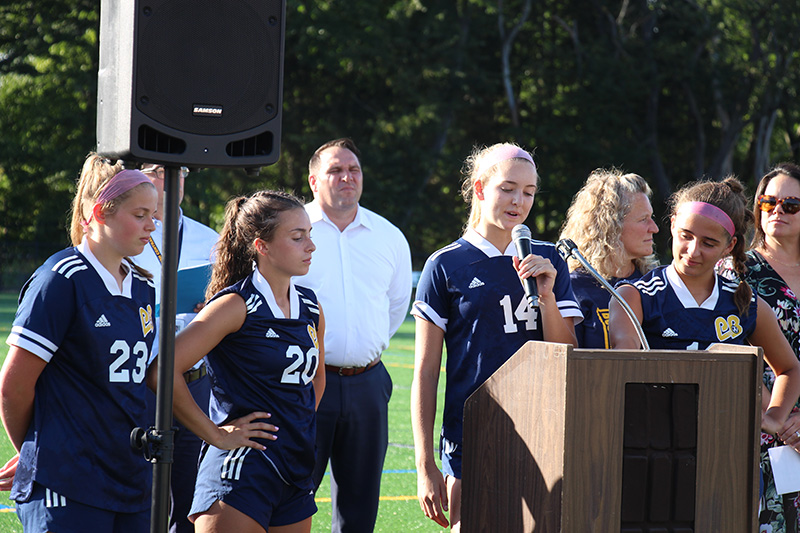 Four high school girls, all dressed in soccer uniforms, stand at a podium where one of them speaks.