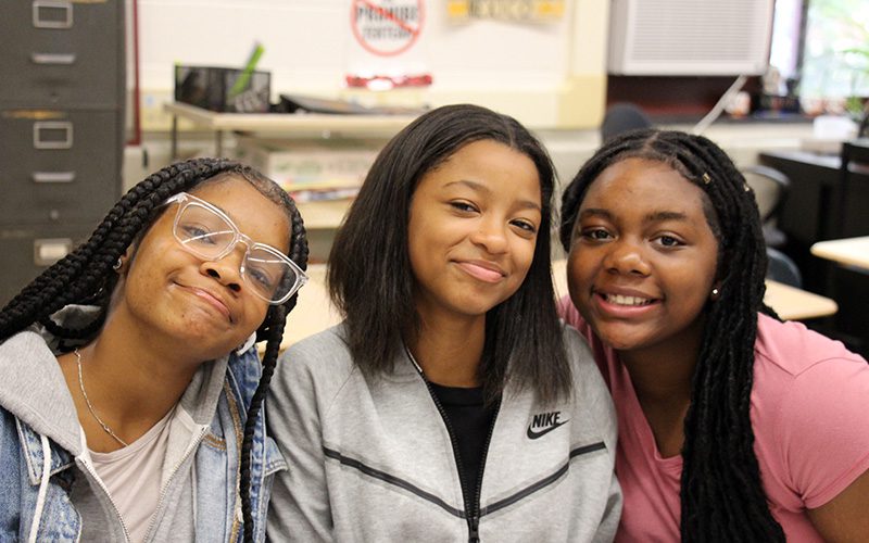 Three high school girls lean in together and smile.