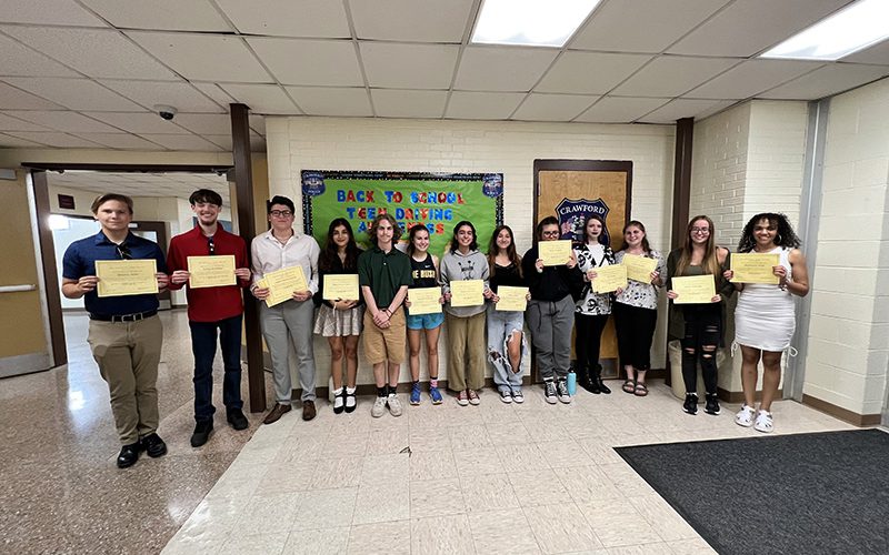 A group of 13 high school students stand in a hallway, all holding award certificates.