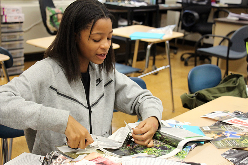 A high school girl cuts pictures out of a magazine as she sits at a table. She is wearing a gray zippered sweatshirt and has shoulder length dark hair.