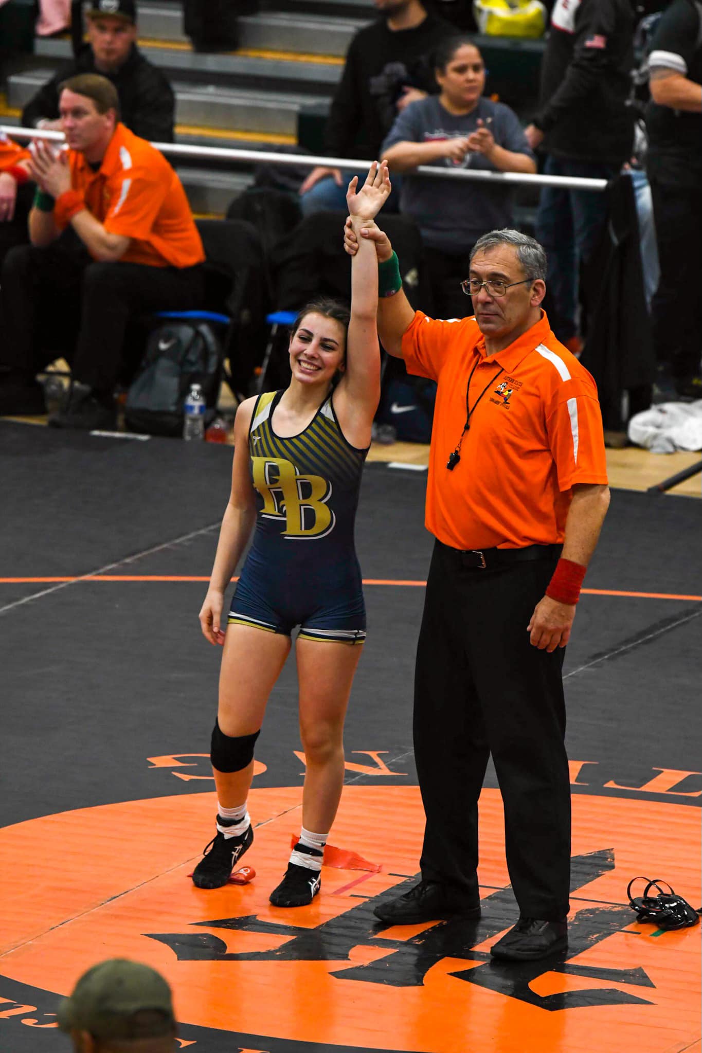 A young woman with dark hair smiles. She is wearing a blue wrestling uniform with a gold PB on the front. A man in an orange shirt holds her hand up high. 