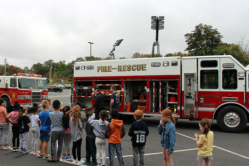 A group of elementary students stand and listen to a firefighter talk to them. Behind him is a red and white fire truck.