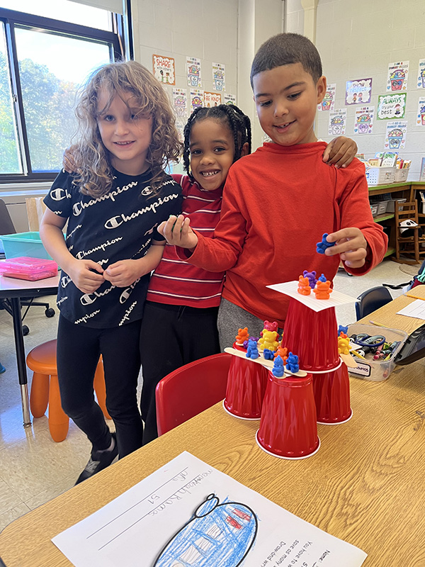 Three first-grade students smile with their STEM project on the table in front of them.