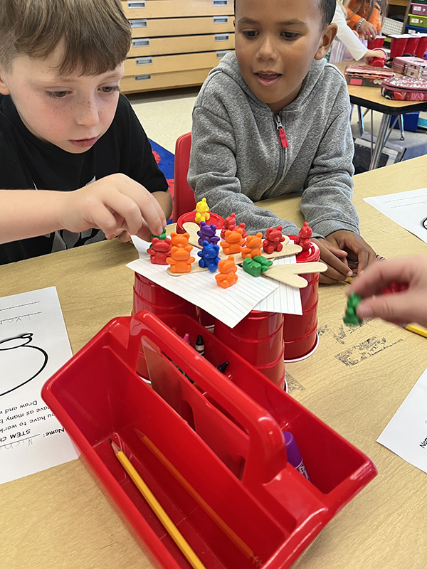 Two first-grade students work on putting little bears on top of cups with sticks and index cards.