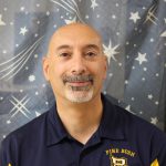 A man smiles. He has a salt and pepper goatee and no hair. He is wearing a blue polo shirt that says Pine Bush on the right side.