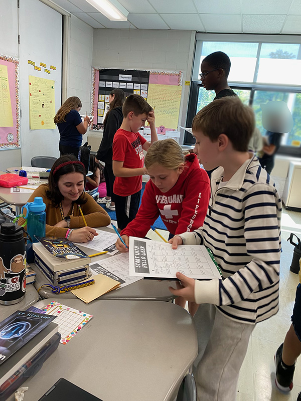 A group of three fifth-grade students work together at a table. Behind them are more students.