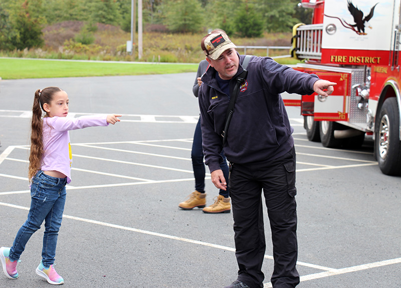 A young elementary student wearing a pink shirt and jeans points to something along with a firerighter.