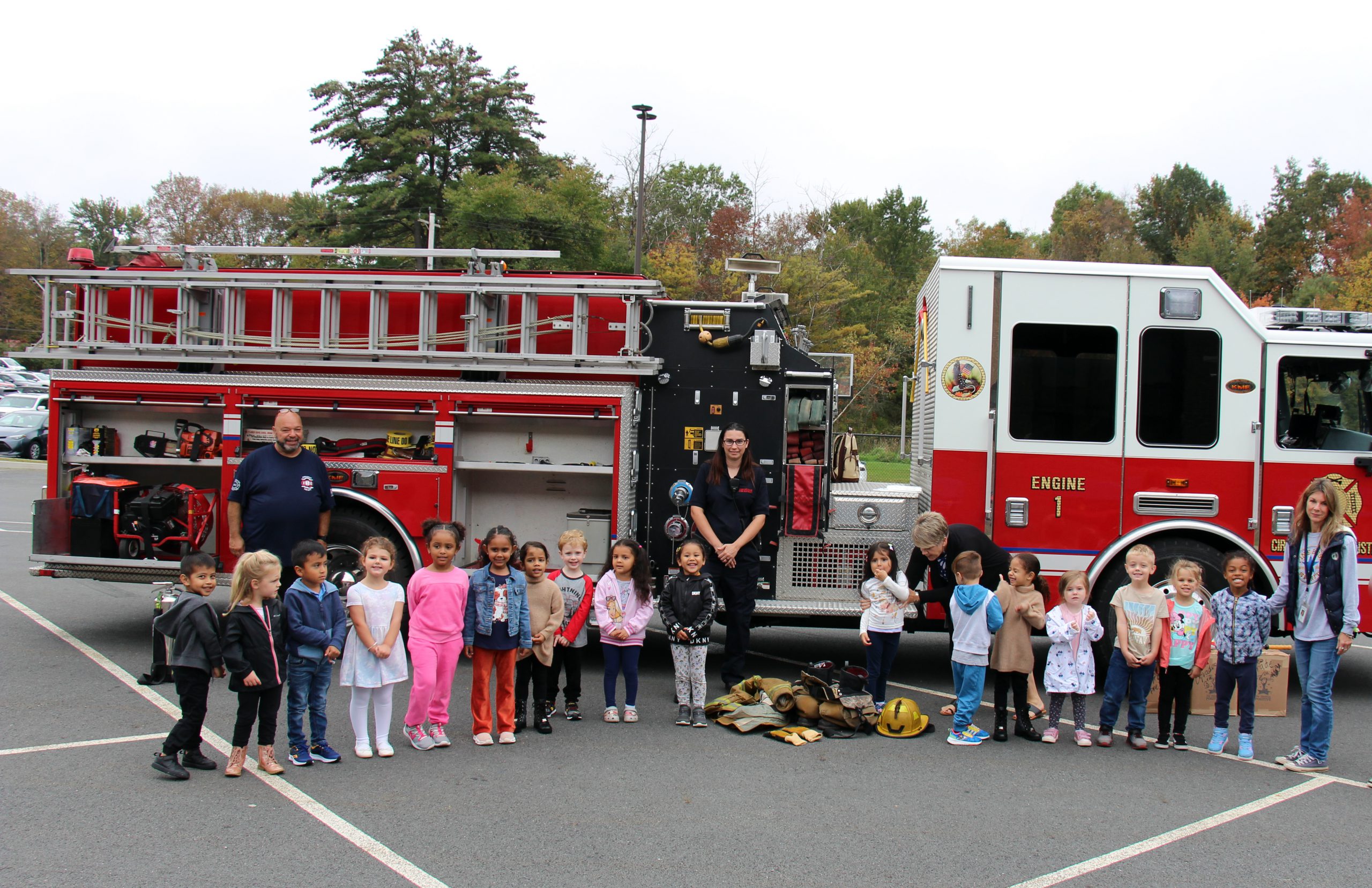 A class of pre-K students stand in front of a fire truck with two firefighters and their teacher.