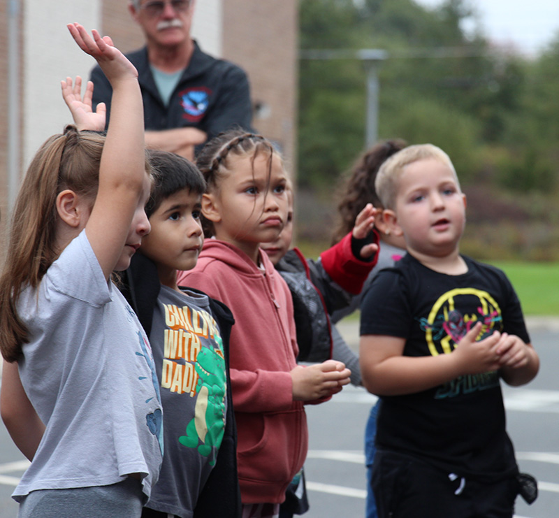 A line of four pre-K students stand listening and one raises her hand.
