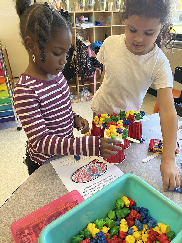 Two first grade girls stand near a table and work on a project balancing little bears on sticks and cups.