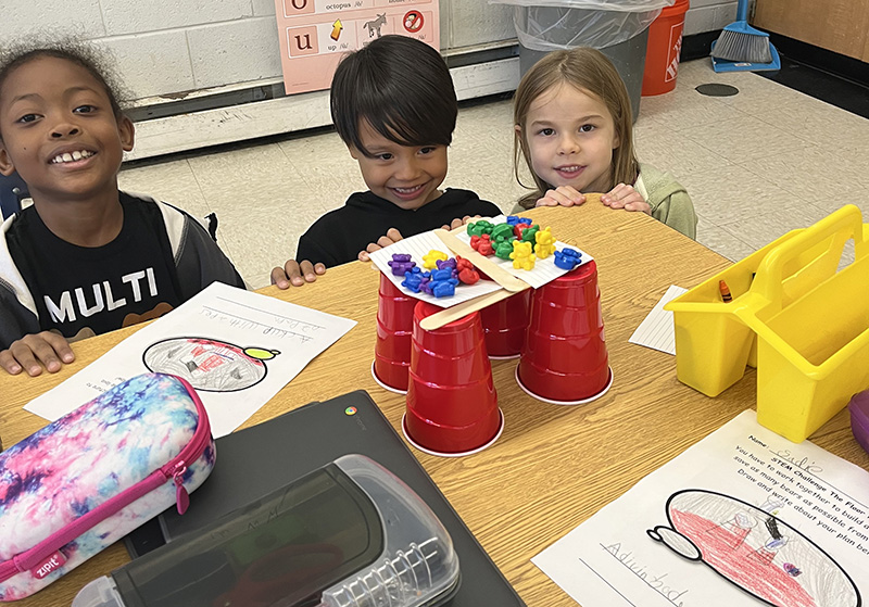 Three first graders smile at their project, which has four cups with sticks on top and index cards with little bears balanced on top.