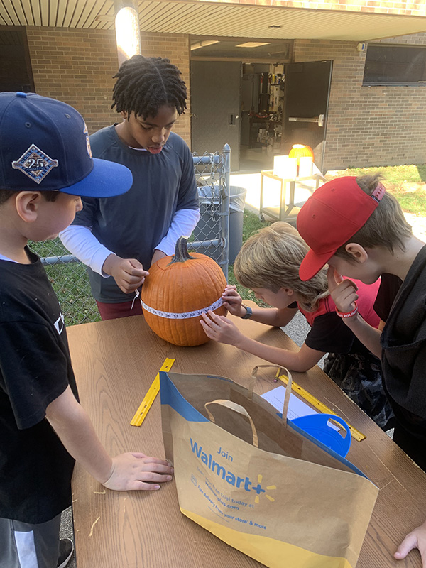 Three middle school students watch as another takes a tape measure and wraps it around a pumpkin.