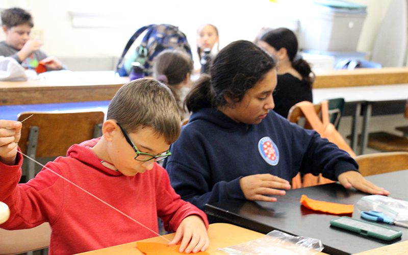 A middle school boy and girl sit at a table and sew on orange pieces of felt.