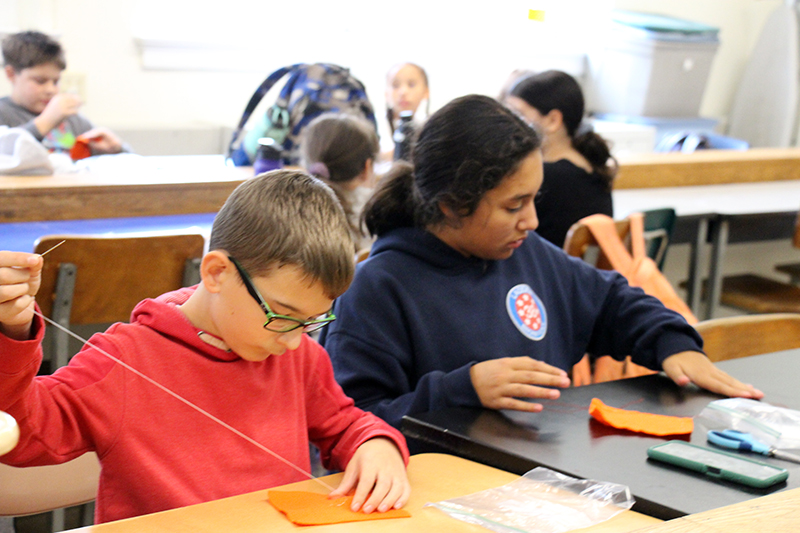 A middle school boy and girl sit at a table and sew on orange pieces of felt.